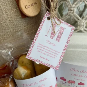 Detalle de la tarjeta con mensaje emotivo para mamá, firmada por Canella en Rama, colgada en el bolso junto al llavero personalizado y con productos del desayuno parcialmente visibles.