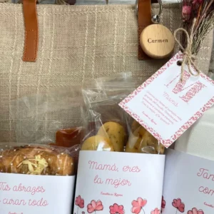 Vista cercana del bolso con productos del desayuno, entre ellos bollería y panecillos envueltos con etiquetas personalizadas con frases como "Mamá, eres la mejor", "Tus abrazos lo curan todo", junto a la tarjeta de felicitación y flores secas.