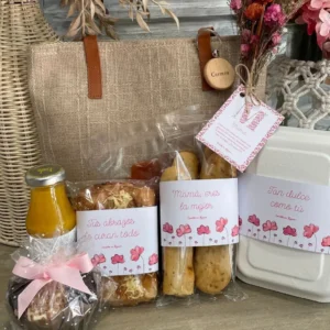 Desayuno completo del Día de la Madre 2025: bolso personalizado con nombre, bollería variada, bebida, muffin con lazo rosa, caja blanca cerrada y etiquetas con mensajes cariñosos sobre mamá.
