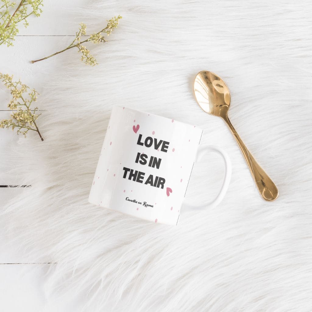 Parte trasera de la taza con la frase "love is in the air" en negro con patrones de corazones, sobre tabla blanca