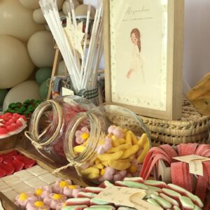 Vista cercana de una mesa dulce con golosinas de distintos tipos: fresas, nubes, gominolas rojas y verdes. Cada detalle está cuidadosamente decorado, incluyendo etiquetas personalizadas y lazos que completan la estética del evento.
