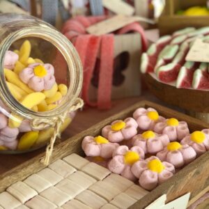 Primer plano de un tarro de cristal lleno de chuches en forma de flores rosas con centro amarillo y plátanos amarillos de azúcar. Las golosinas están organizadas en bandejas de madera para un toque rústico y elegante.