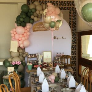Montaje de mesa dulce decorada con un estilo delicado y natural, combinando tonos beige, verde y rosa pastel. Incluye globos, flores naturales, tarros de chuches personalizadas y carteles con diseño ilustrado.