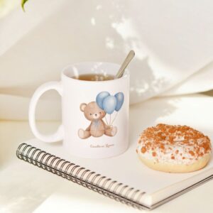 Taza blanca con ilustración de osito y globos azules, junto a un cuaderno, una rosquilla y una taza de té