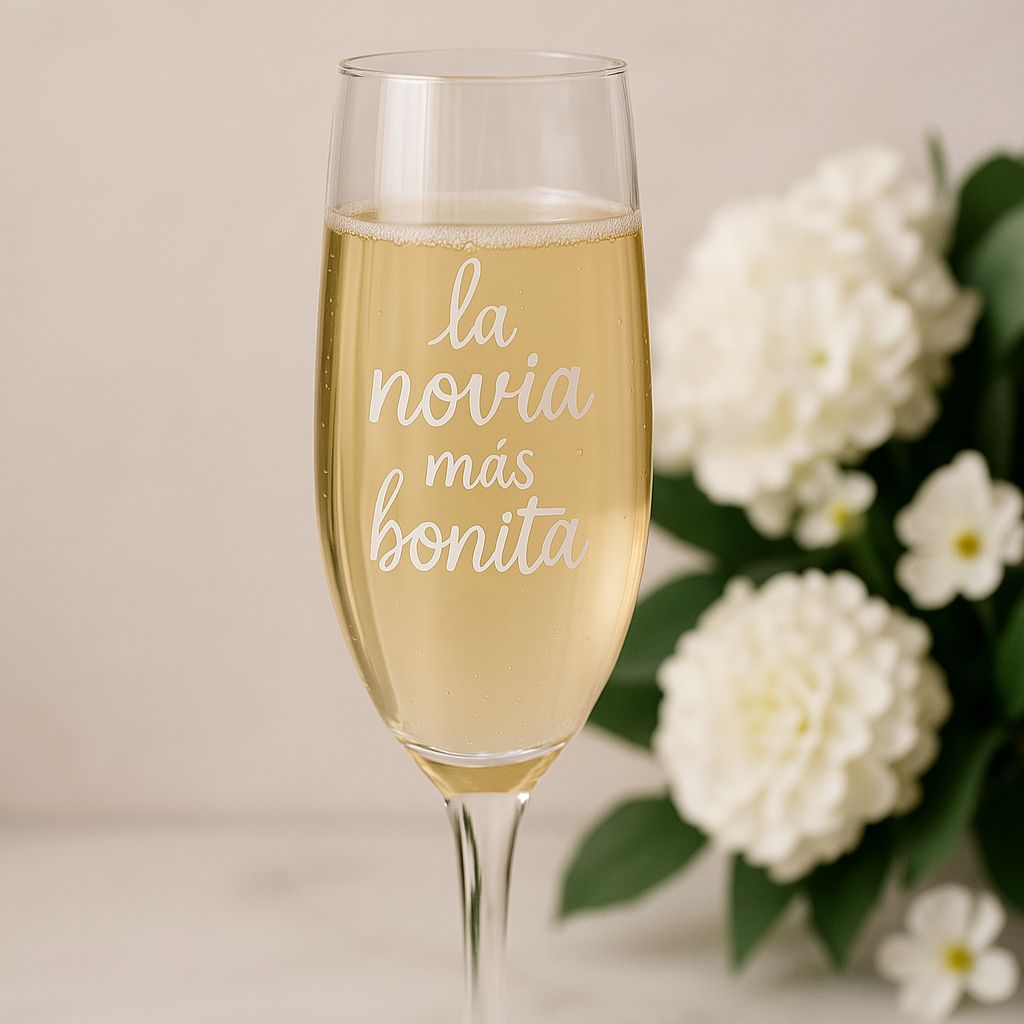 Copa de cava para novia con la frase “la novia más bonita” grabada, decorada con flores blancas al fondo.