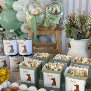 Primer plano de recipientes con palomitas, latas y botes de chuches decorados con ilustraciones de perro salchicha para mesa dulce de comunión.