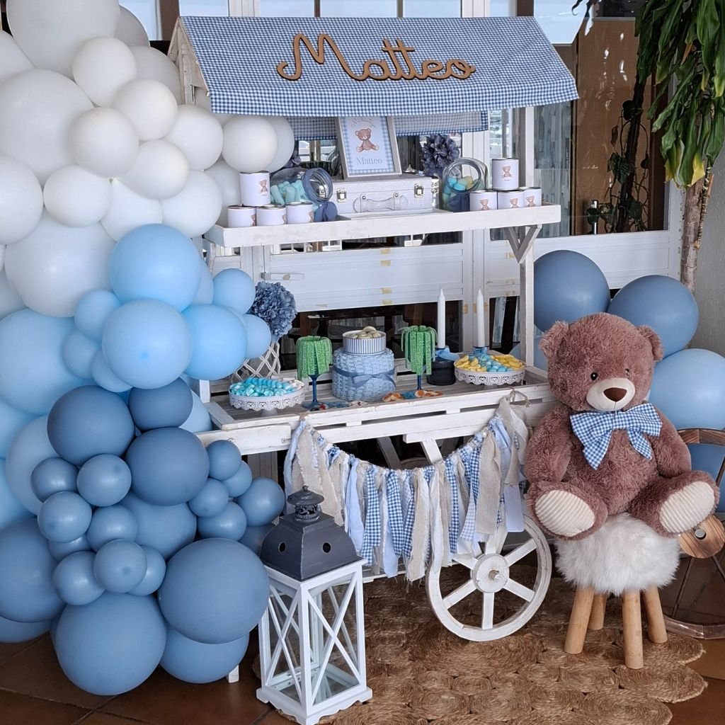 Carrito de golosinas para bautizo decorado con globos azul pastel, peluche y dulces variados.