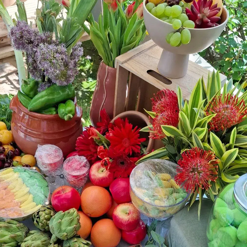 Detalle de frutas tropicales como alcachofas, limones, naranjas y manzanas junto a flores rojas y recipientes decorativos